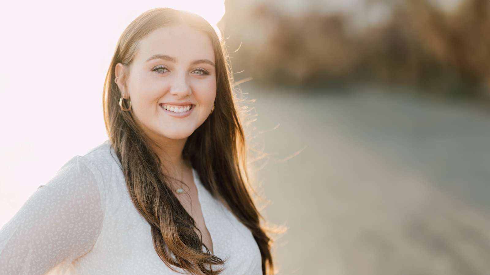 Young woman with long brown hair smiling, wearing a white top, standing outdoors with sunlight behind her.