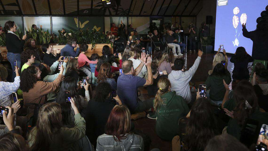 Audience sitting and raising hands, many holding up phones, facing a screen showing coral reef images.