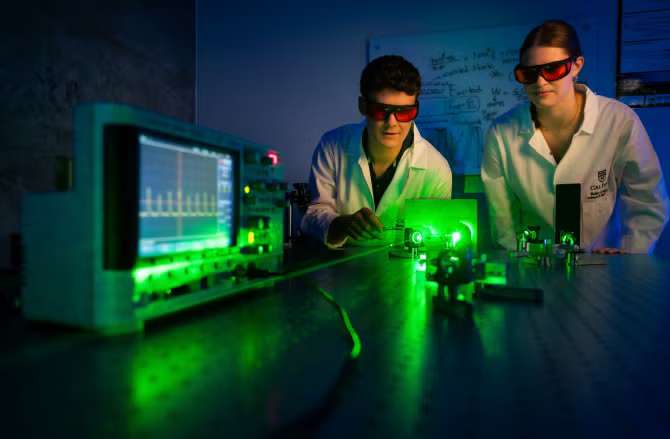 Two scientists in lab coats and safety glasses working with green laser equipment and an oscilloscope.
