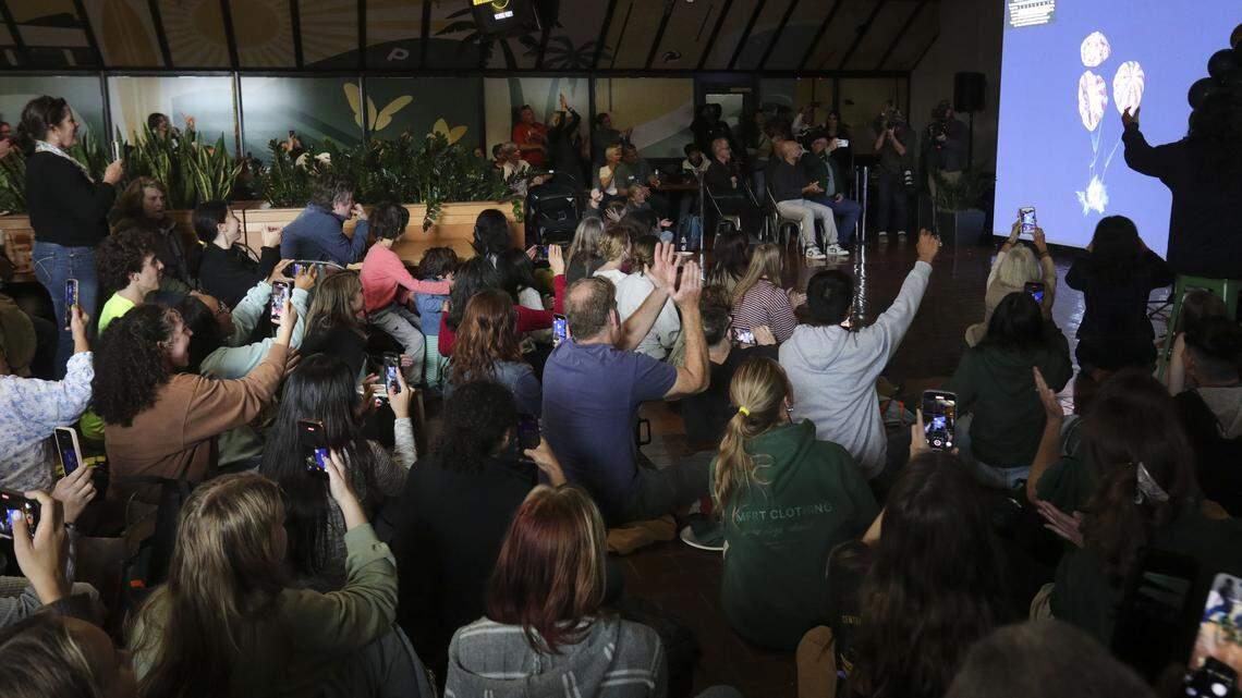 Audience sitting and raising hands, many holding up phones, facing a screen showing coral reef images.
