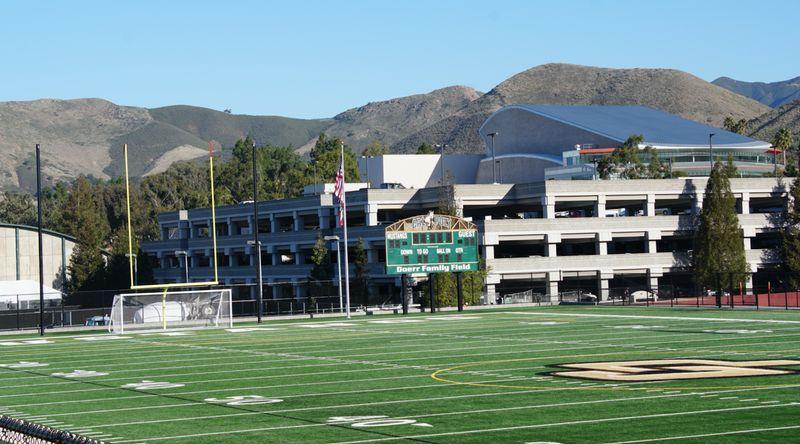 Football field with scoreboard reading "Doerr Family Field," goalpost, and parking garage with hills in the background.