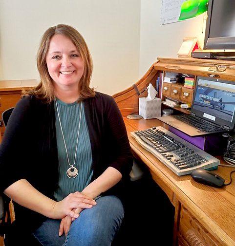 Woman with shoulder-length hair smiling, seated at wooden desk with computer and keyboard.