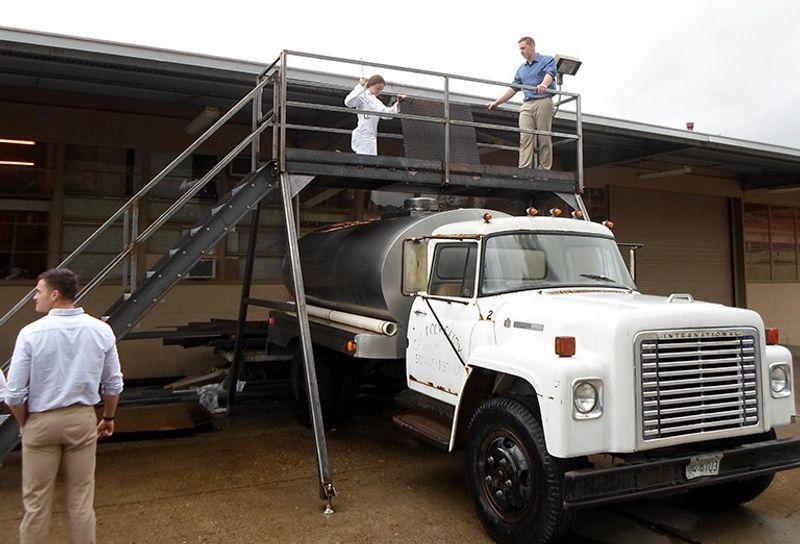 White industrial tanker truck with two people on an elevated metal platform above it and one person standing nearby.