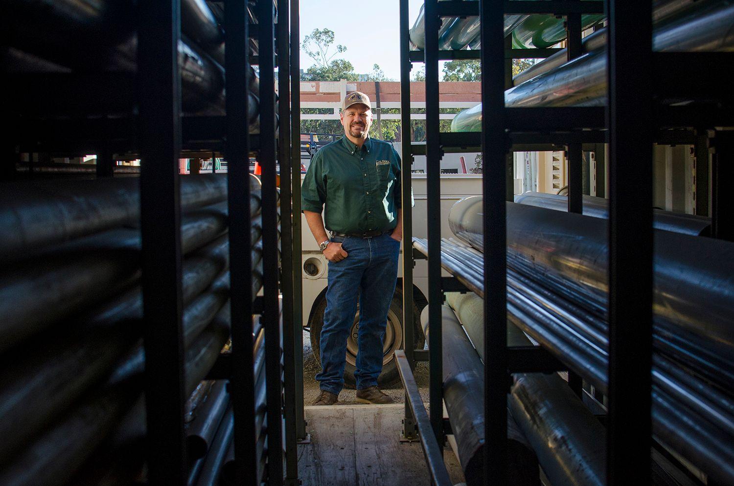 Man in green shirt and cap standing between metal pipes stored on racks outdoors.