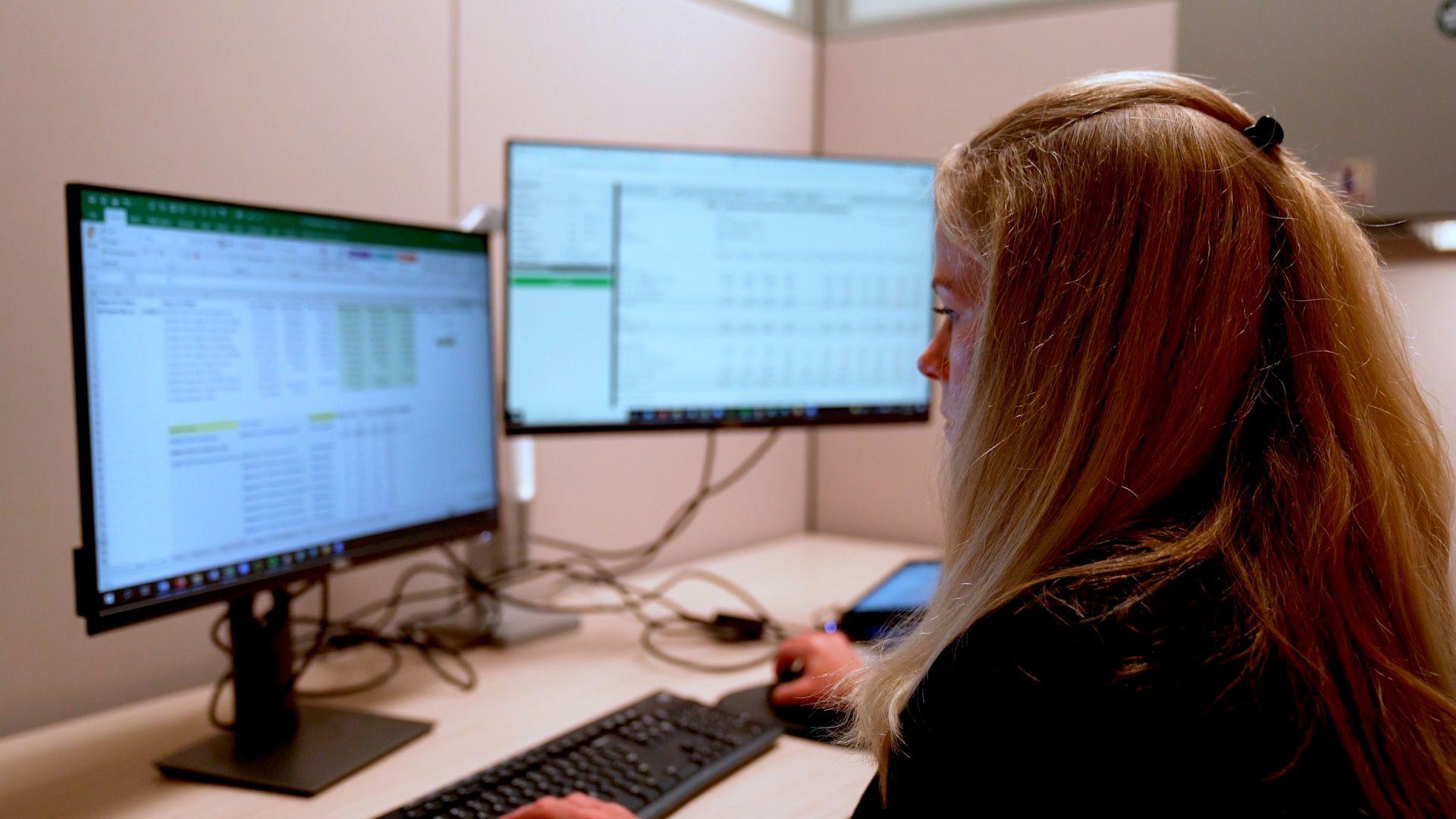 Woman with long hair working at desk with two computer monitors showing spreadsheets and data.