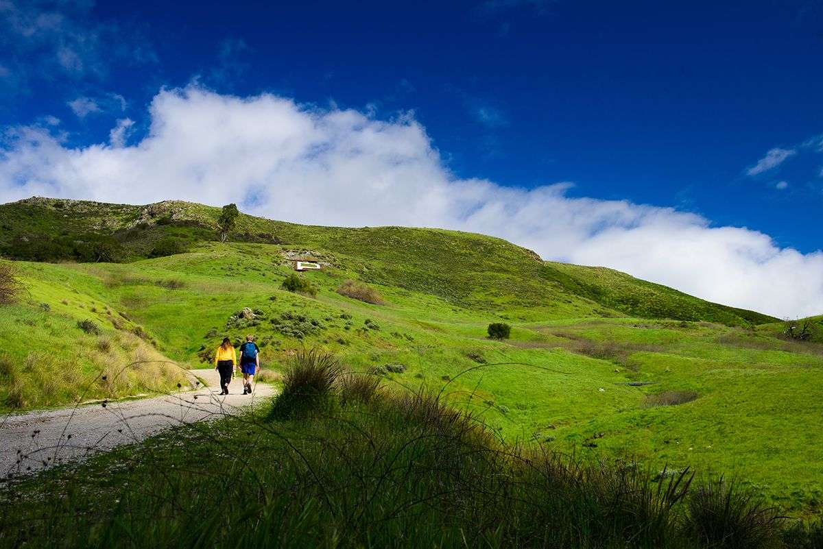 Two people walking on a dirt path in a lush green hilly landscape under a blue sky with scattered clouds.
