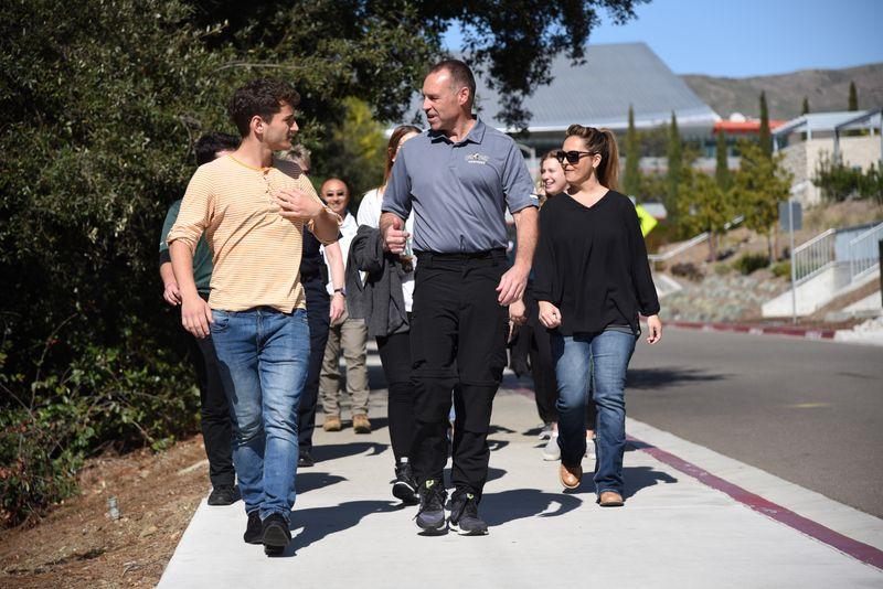 Group of adults walking and talking on a sunny sidewalk lined with trees and buildings.