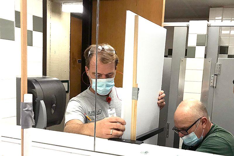 Two masked men setting up a white partition in a public restroom with tiled stalls in the background.