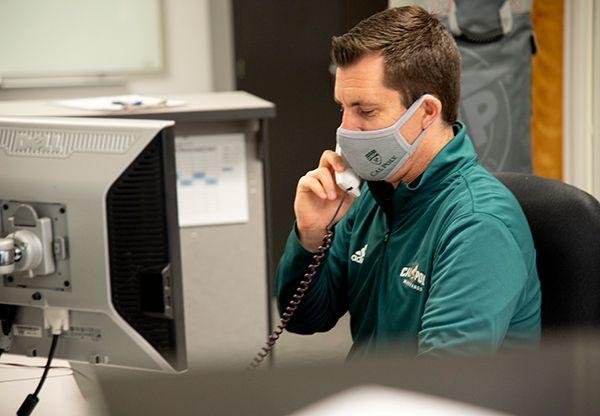 Man wearing a face mask and green jacket using a corded phone at an office desk with a computer monitor.