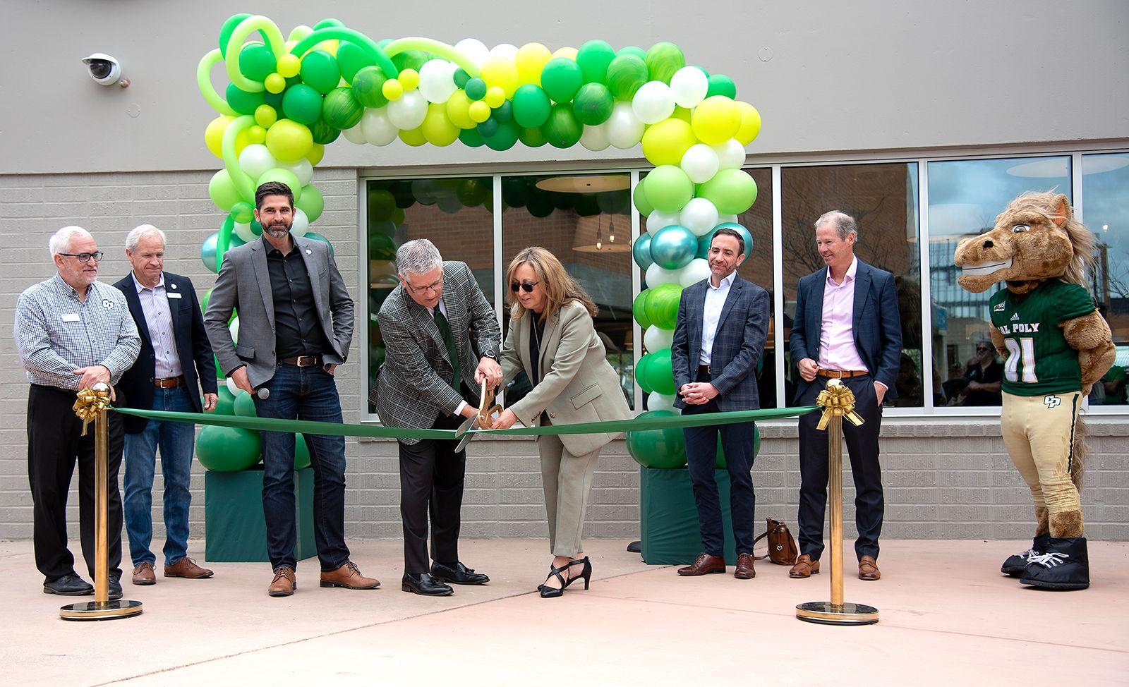 People at a ribbon-cutting ceremony with a green, yellow, and white balloon arch and a mascot in a green jersey.