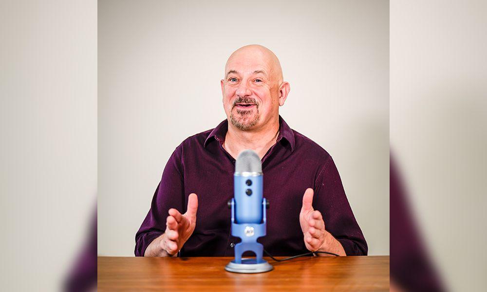 Bald man with goatee in purple shirt speaking and gesturing behind a blue microphone at a wooden table.