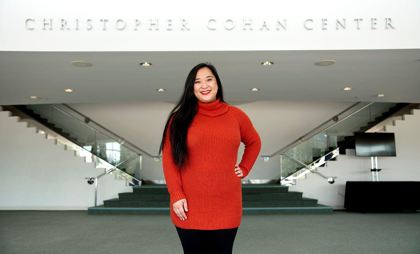 Woman with long black hair in a red sweater standing in front of stairs under a sign reading "Christopher Cohan Center