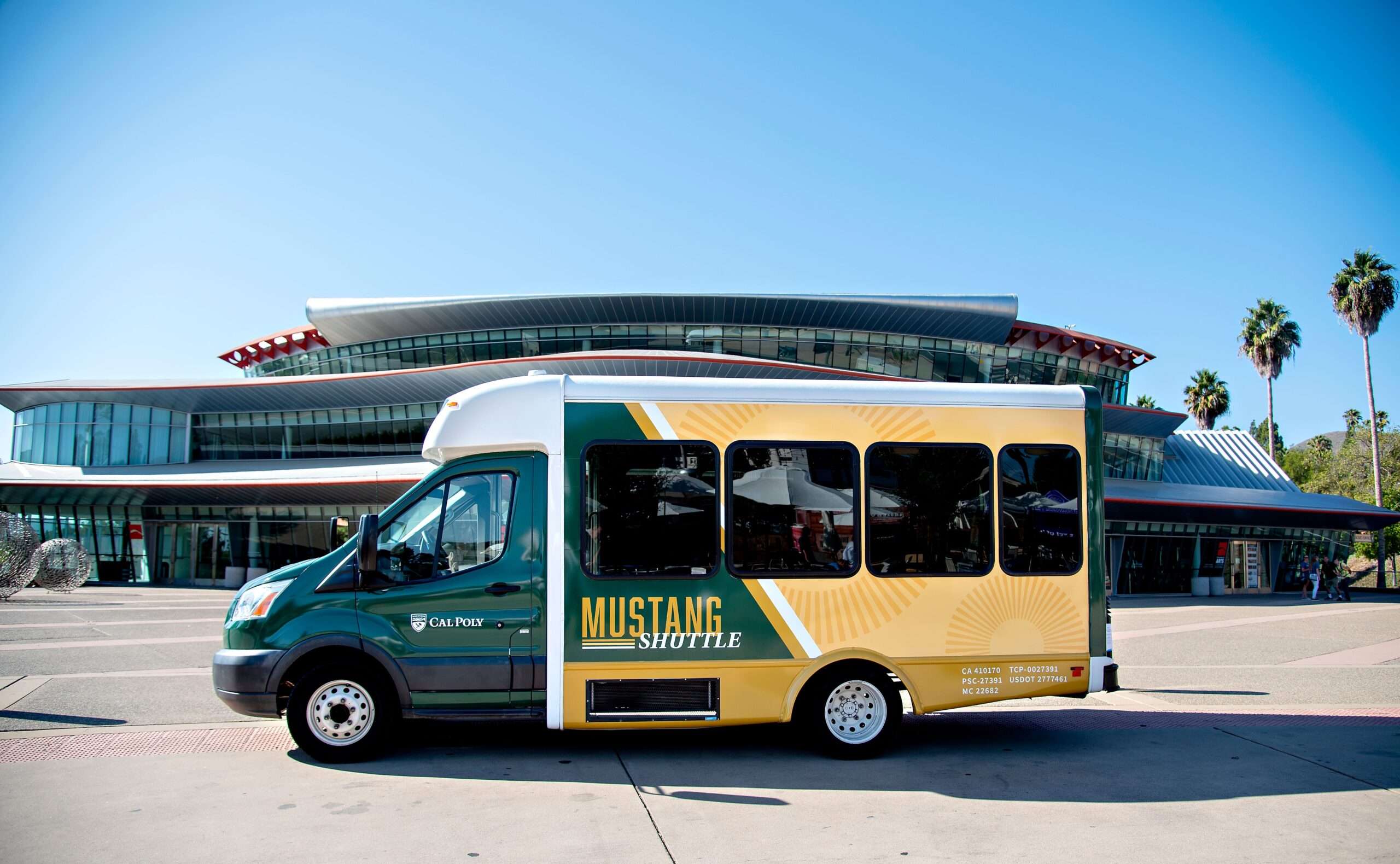 Cal Poly Mustang Shuttle bus parked in front of a modern glass building with palm trees in the background.