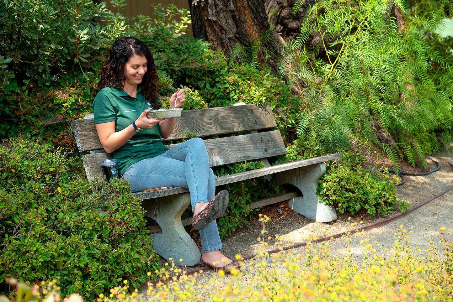 Woman in green shirt and jeans sitting on a wooden bench eating from a foil container in a garden area.