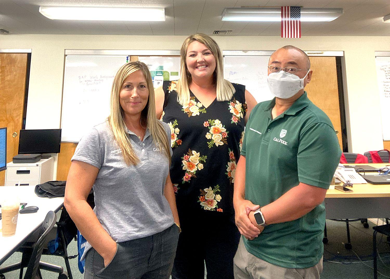 Three people stand in an office, two wearing Cal Poly shirts, one man is wearing a face mask and smartwatch.