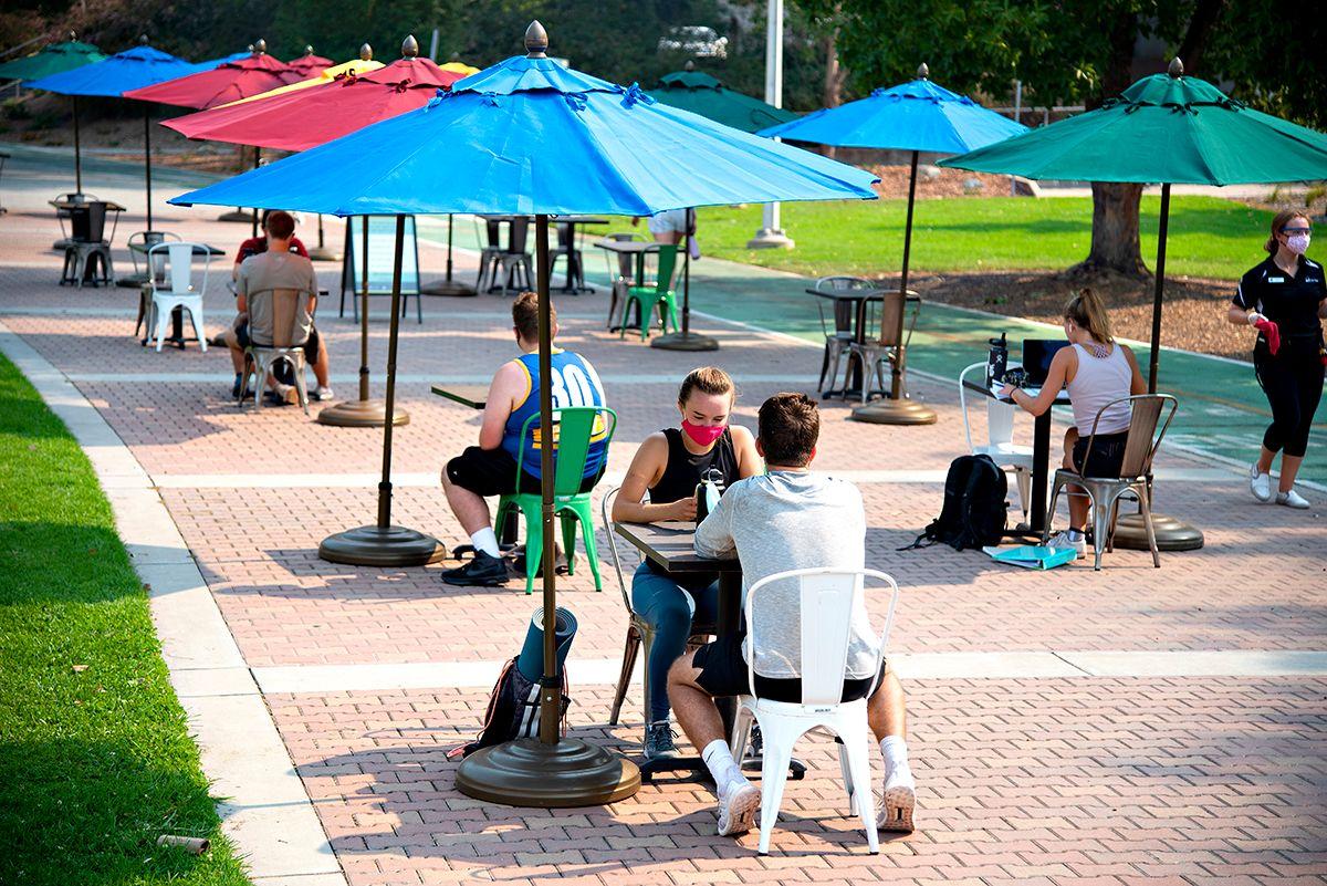 Outdoor seating with colorful umbrellas and people sitting in pairs, some wearing masks, on a paved pathway.