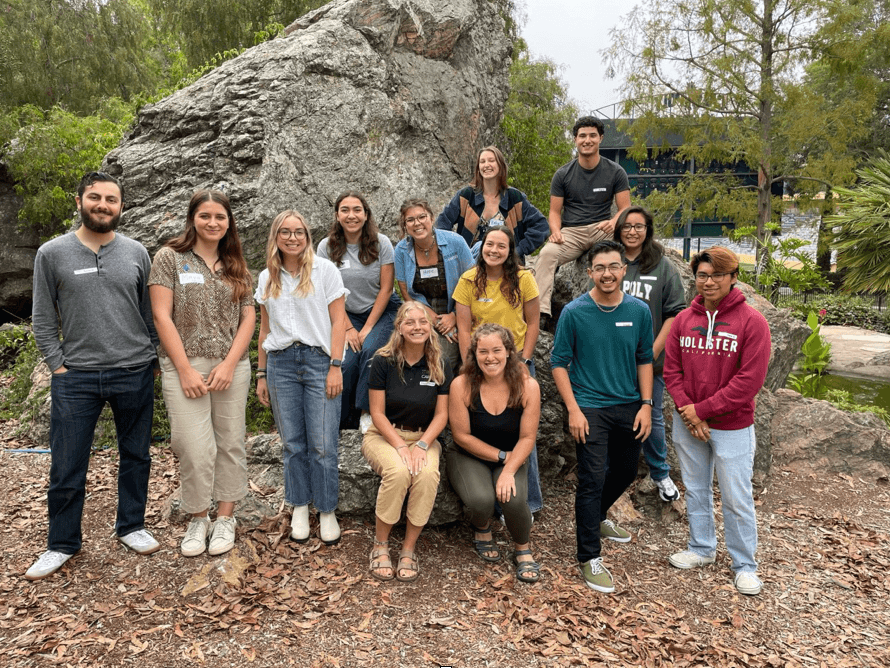 Thirteen young adults posing outdoors in front of a large rock and trees, smiling at the camera.