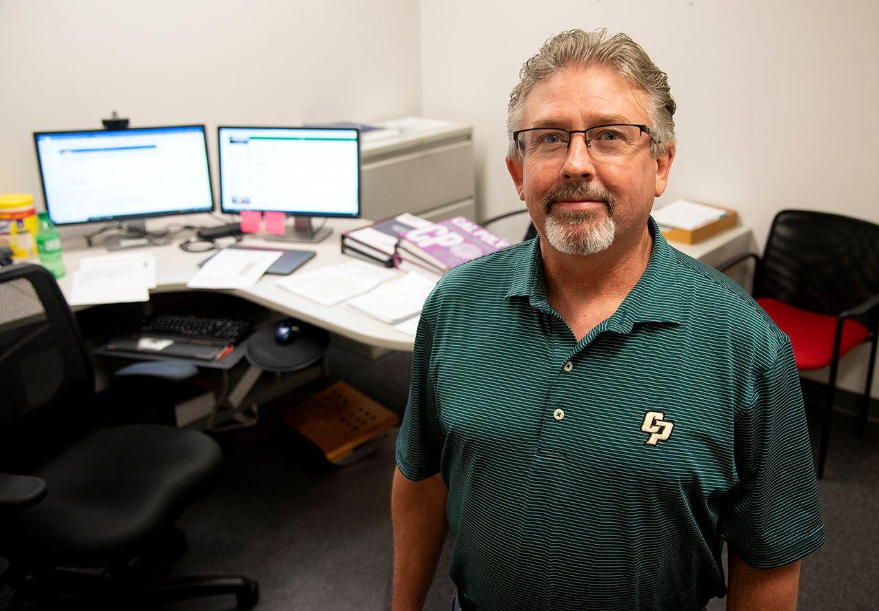 Man with glasses and salt-and-pepper hair wearing a green striped polo shirt standing in an office with dual monitors and papers.