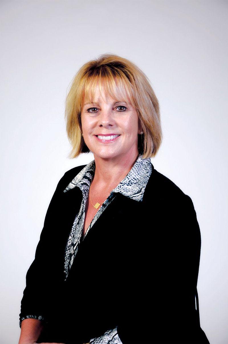 Smiling middle-aged woman with short blonde hair wearing a black blazer and snakeskin-patterned shirt against a white background.