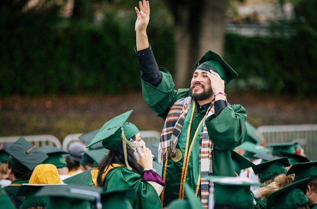 Graduate in green cap and gown waving to crowd among fellow graduates outdoors.