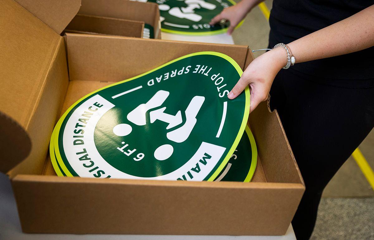 Person holding a green circular sign that reads "Maintain 6 ft physical distance STOP THE SPREAD" from an open cardboard box.