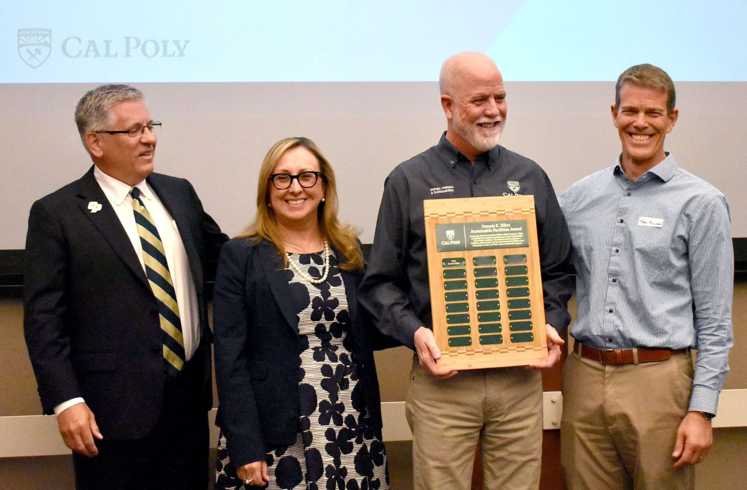 Four people smiling, one holding a wooden plaque labeled "Dennis K. Elliot Sustainable Facilities Award" with Cal Poly logo.