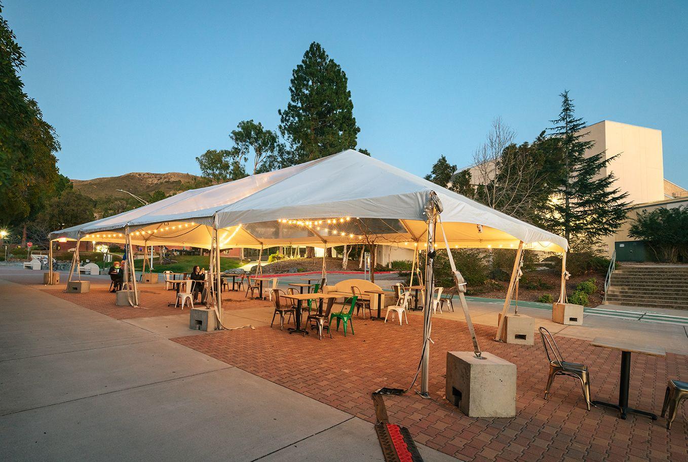Outdoor seating area under a white tent with string lights, tables, chairs, and a few people seated during dusk.