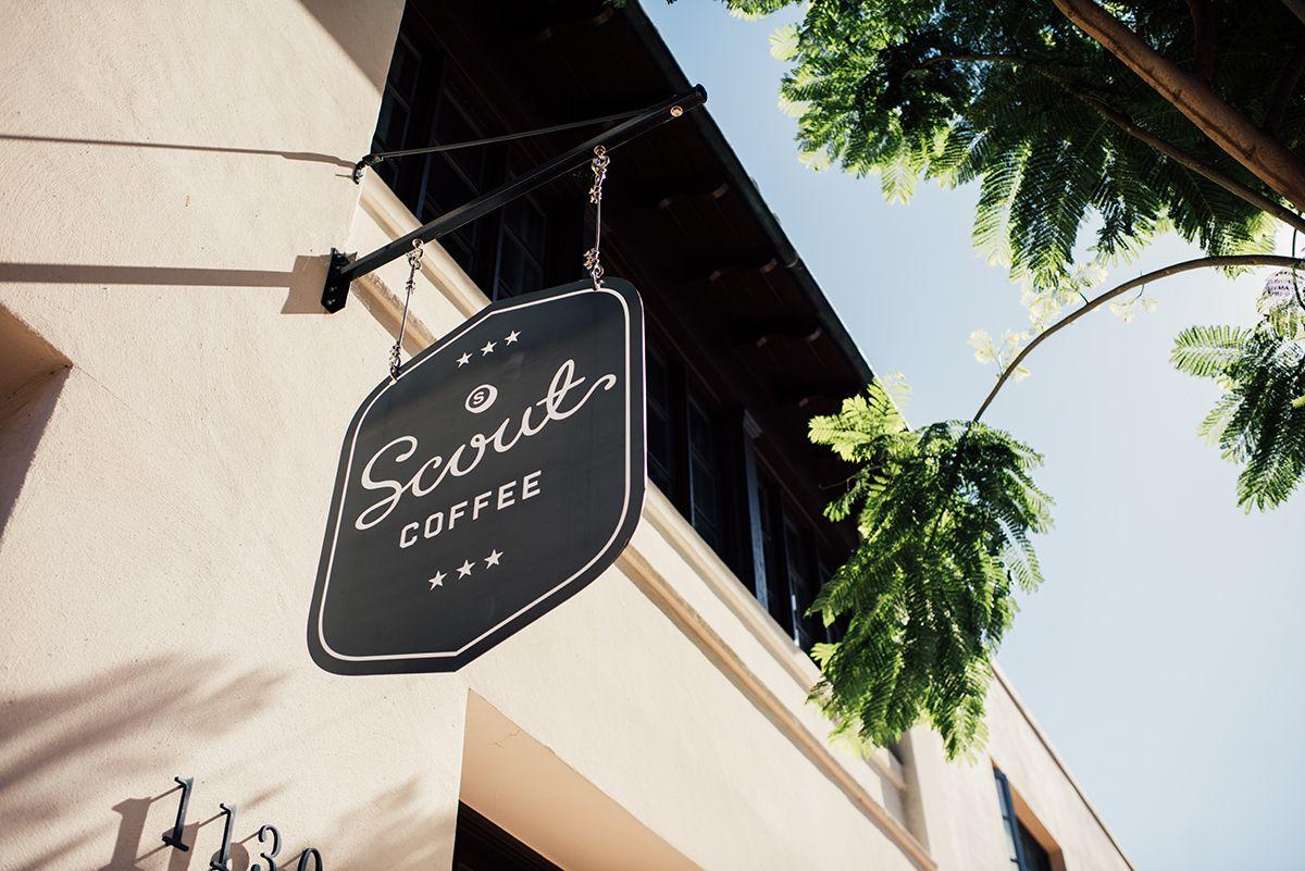 Black hanging sign with white text "Scout Coffee" outside a beige building with trees above.