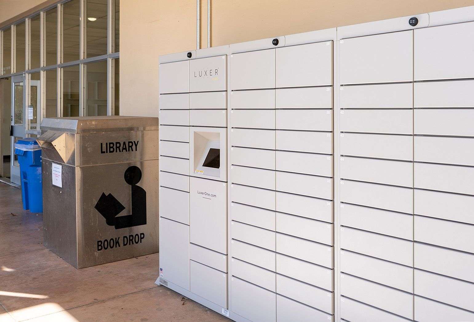 Library book drop bin next to a modern parcel locker with multiple compartments and a touchscreen panel.
