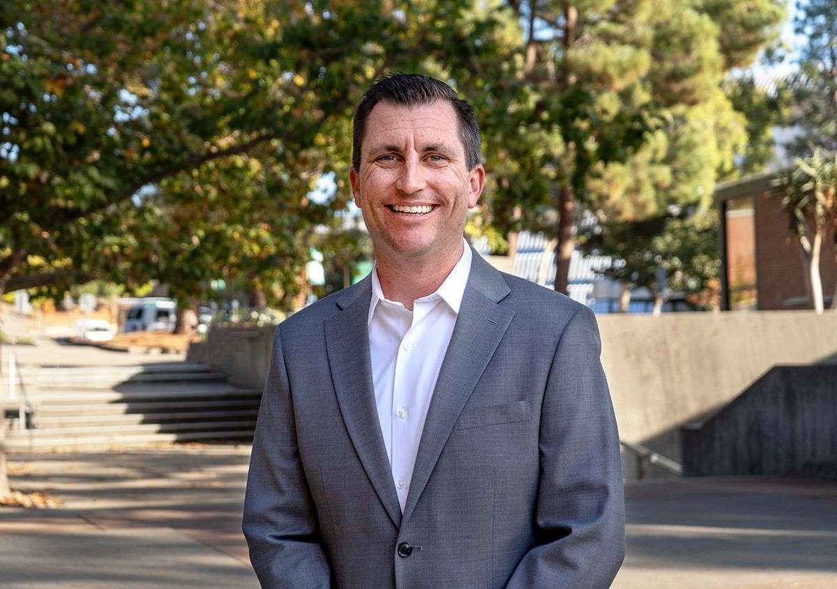 Man in gray suit jacket and white shirt smiling outdoors with trees and steps in the background.