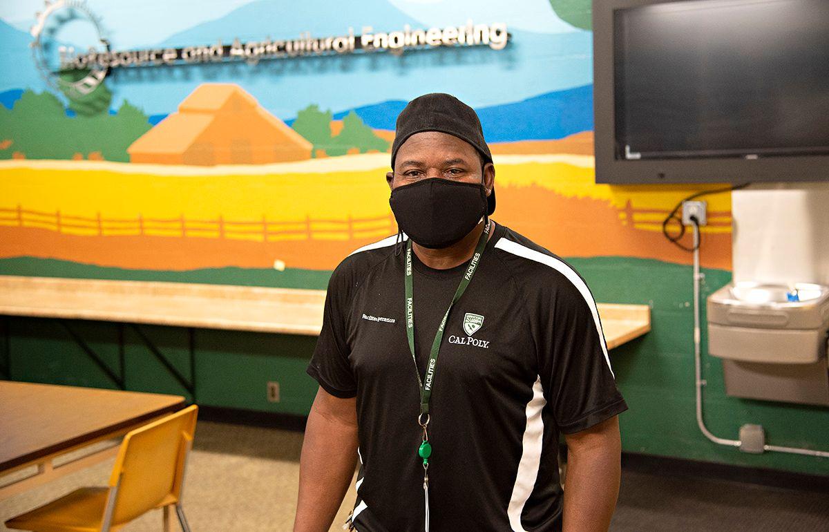Man wearing black mask and Cal Poly shirt standing inside room with mural and TV on wall.
