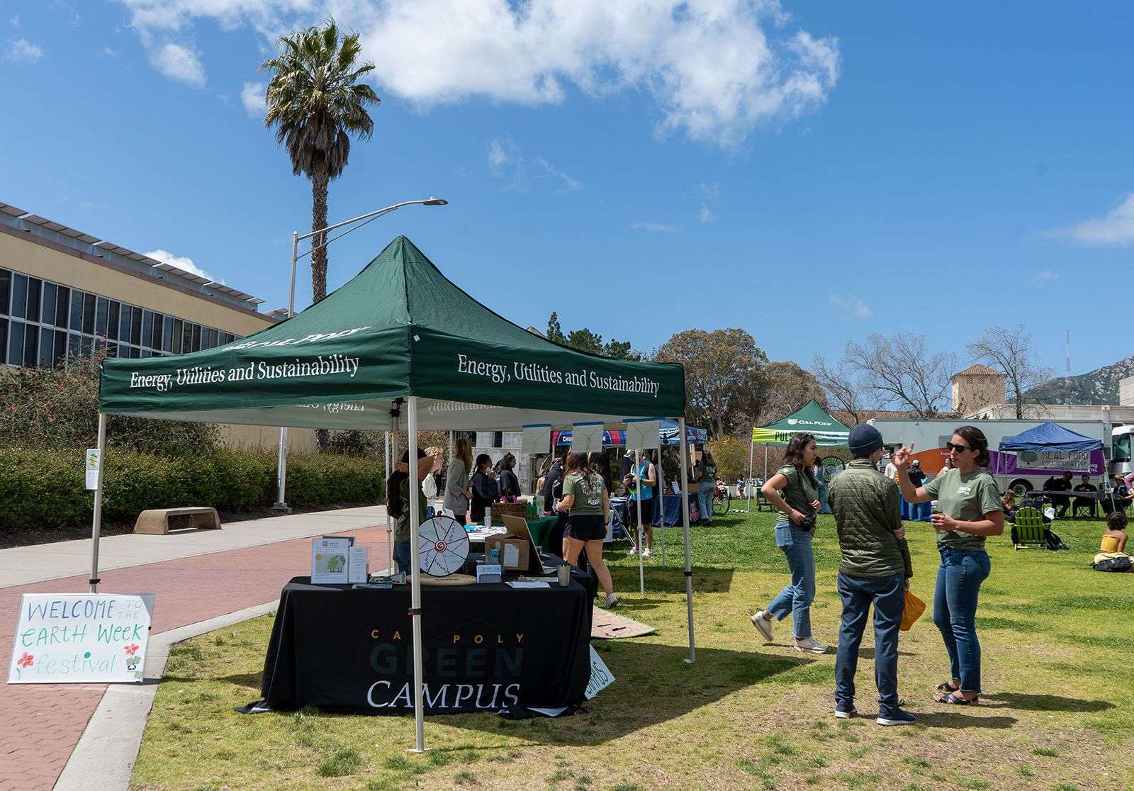 Green tent with "Energy, Utilities and Sustainability" and "Cal Poly Green Campus" with people at an outdoor Earth Week festival.