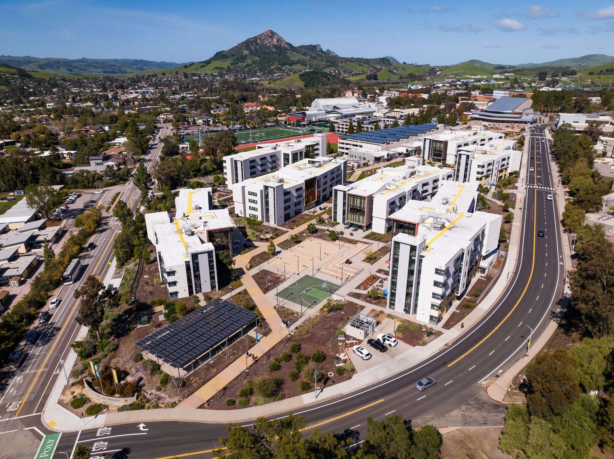 Modern apartment complex with solar panel roofs, sports fields, and surrounding roads in a suburban area with hills in the background.