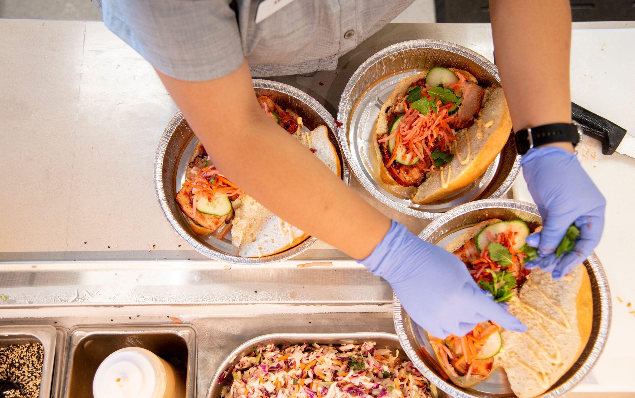 Hands wearing blue gloves preparing sandwiches with grilled meat, cucumber, shredded carrots, and greens in aluminum trays.