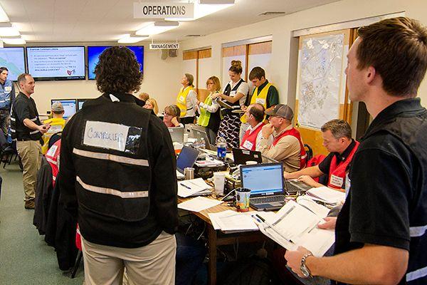 Team of emergency responders and coordinators in a command center with laptops, documents, and screens displaying operations info.