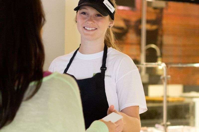 Smiling food service worker wearing visor and apron hands a card to a customer at a counter.