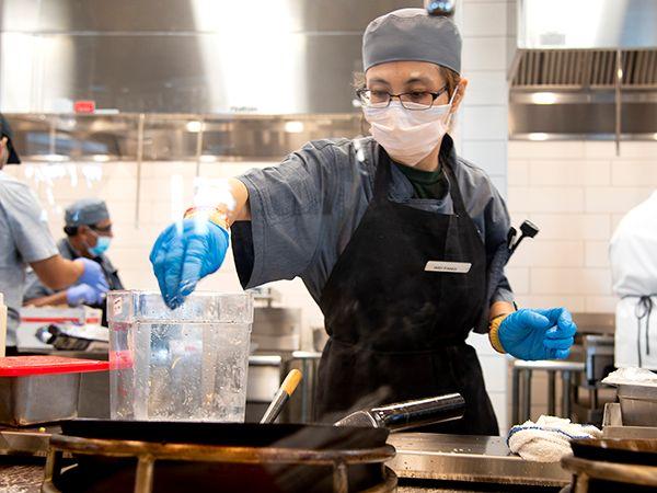 Chef wearing mask, gloves, and apron preparing food in a professional kitchen near a water container and stove.