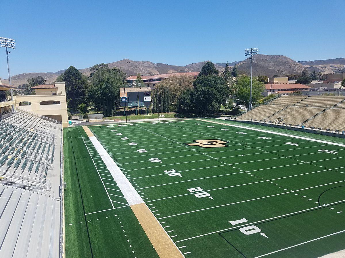 Football field with a large "S" logo at midfield, empty bleachers, scoreboard, and mountainous background under clear sky.
