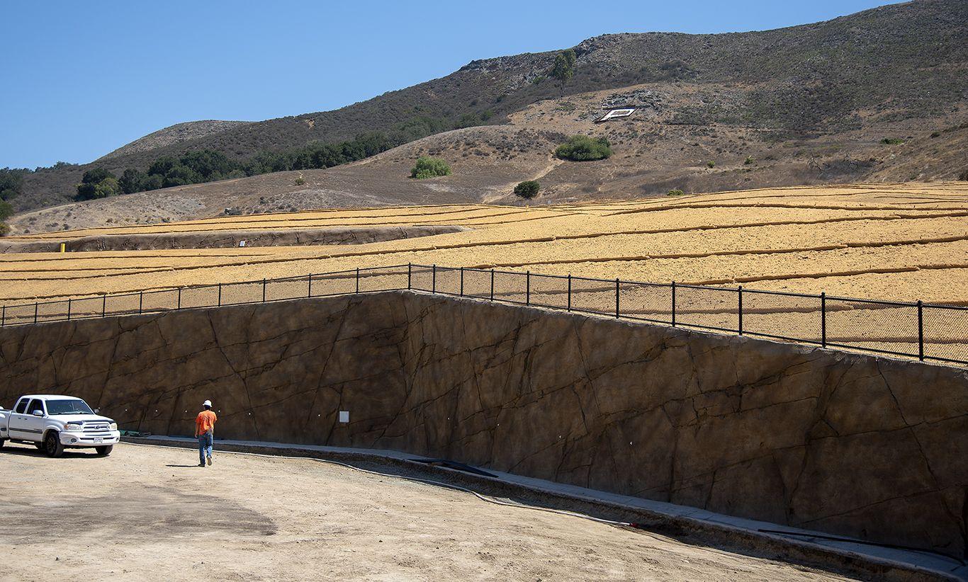 Man in orange shirt walking on dirt near white pickup truck, retaining wall, and terraced dry hillside under clear sky.