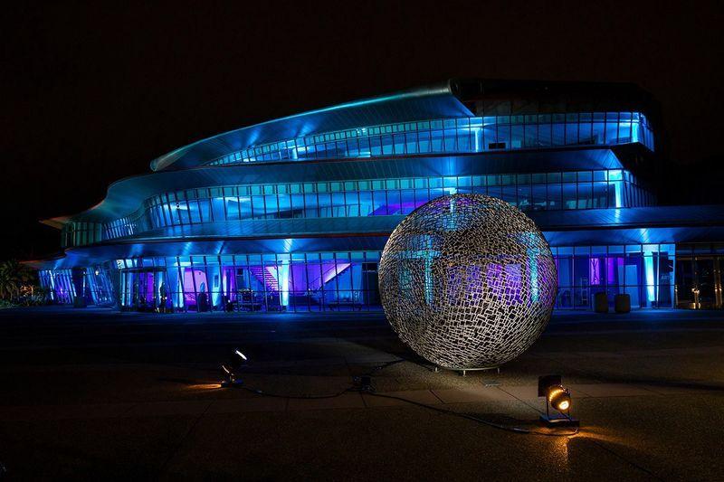 Modern building illuminated in blue and purple lights with a lit spherical metal sculpture in the foreground at night.