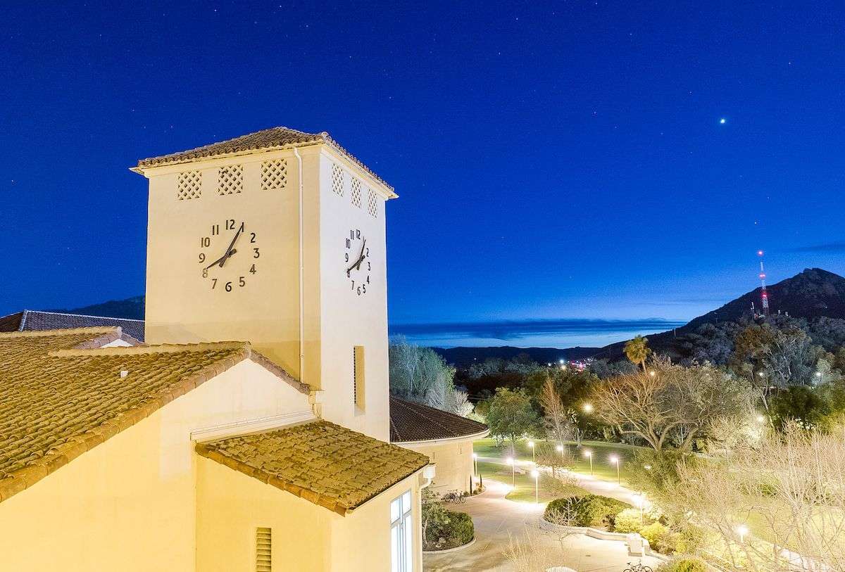 Clock tower on a building at night with illuminated pathways, trees, and a mountain with a red light tower in the background.