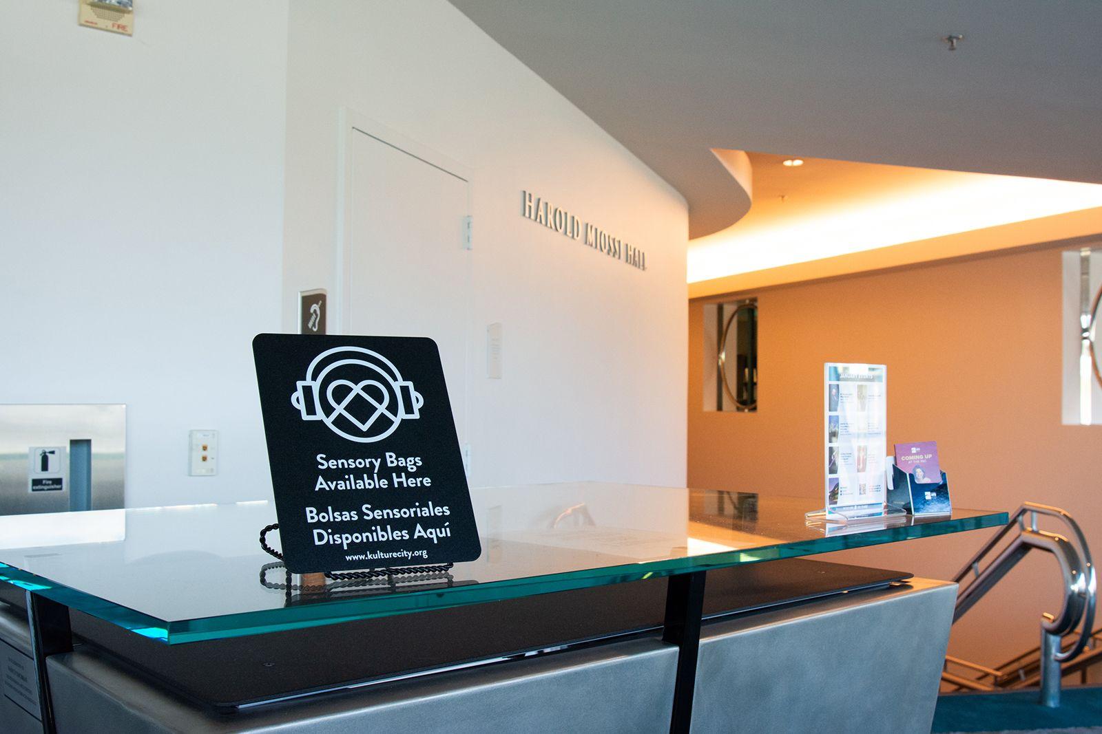 Sign on counter reading "Sensory Bags Available Here / Bolsas Sensoriales Disponibles Aquí" in front of Harold Moss Hall.