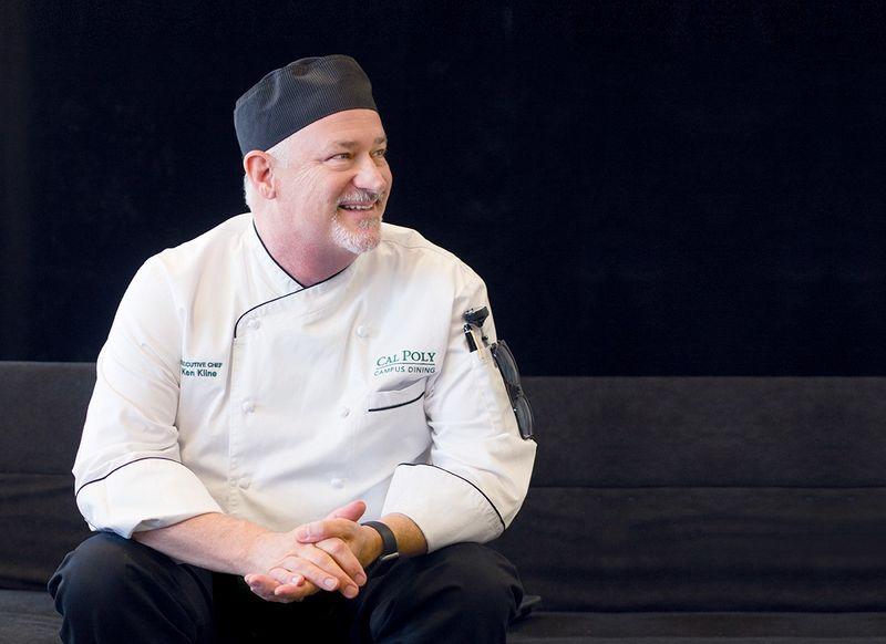 Man in a Cal Poly Campus Dining chef uniform and black hat smiling while sitting on a dark couch.