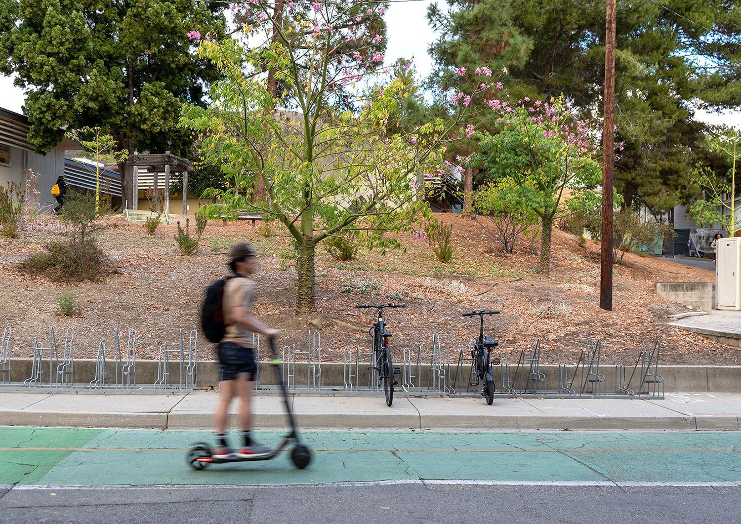 Person wearing a backpack rides a scooter past a bike rack with two parked bicycles and trees in the background.