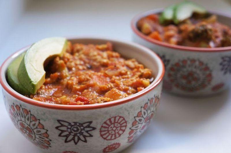 Two bowls of tomato-based stew topped with sliced avocado, in patterned bowls on a white surface.