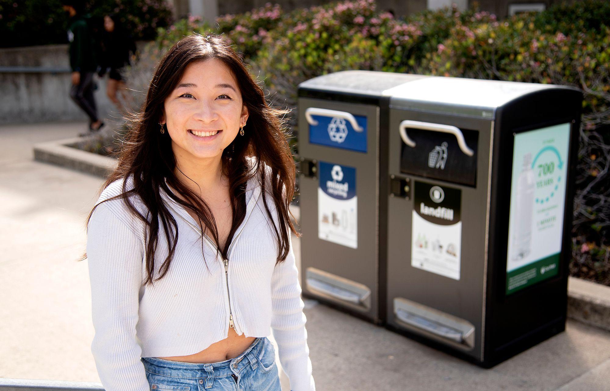Young woman smiling outdoors near paired recycling and landfill bins.