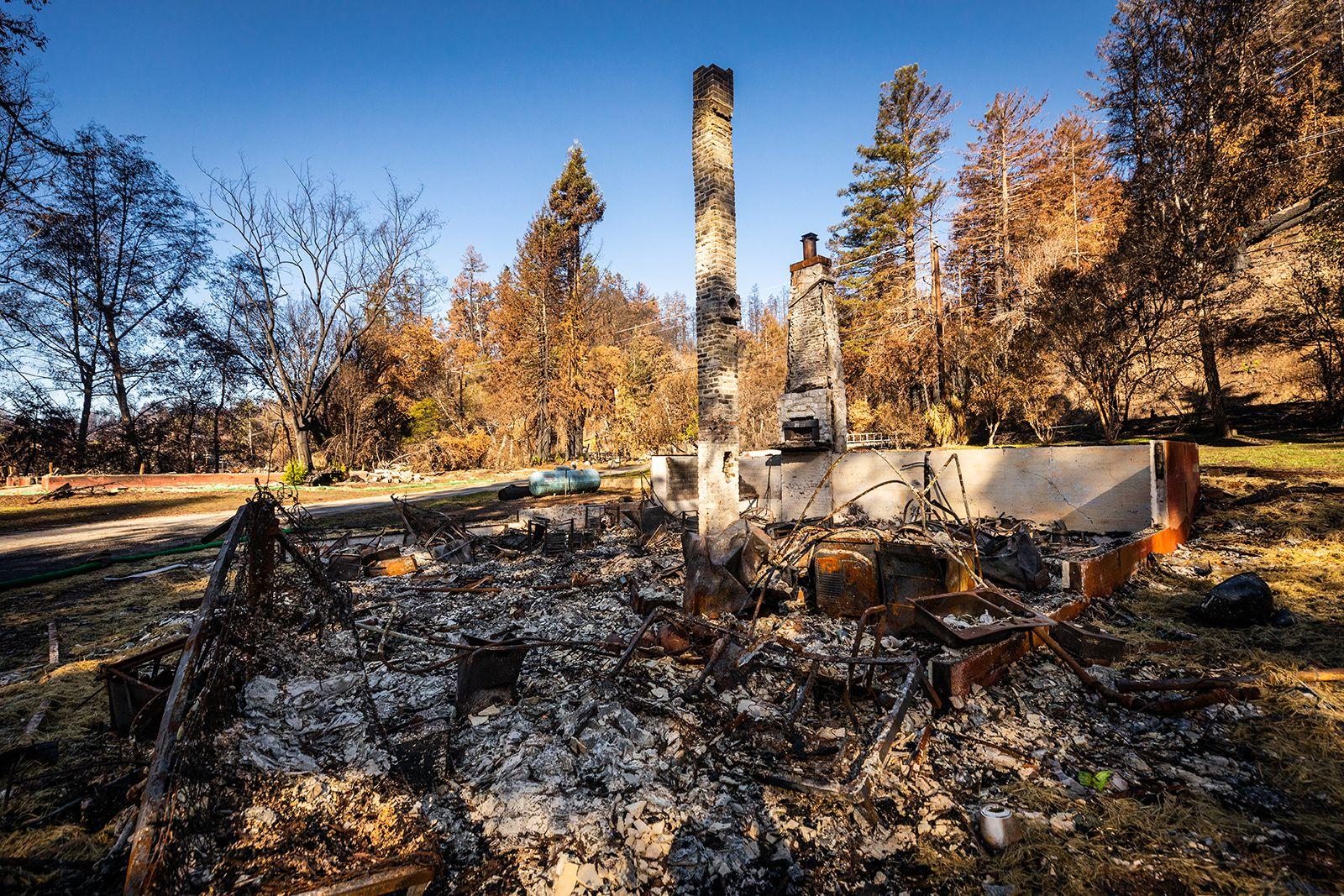 Charred remains and stone chimney of a house destroyed by fire, surrounded by burnt trees under a clear sky.