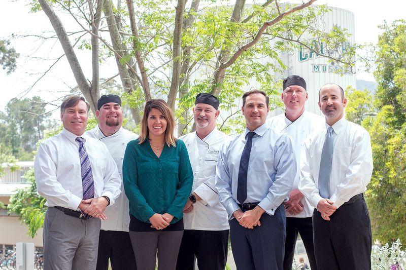 Seven professionals including chefs and office staff stand outdoors with trees and a building in the background.