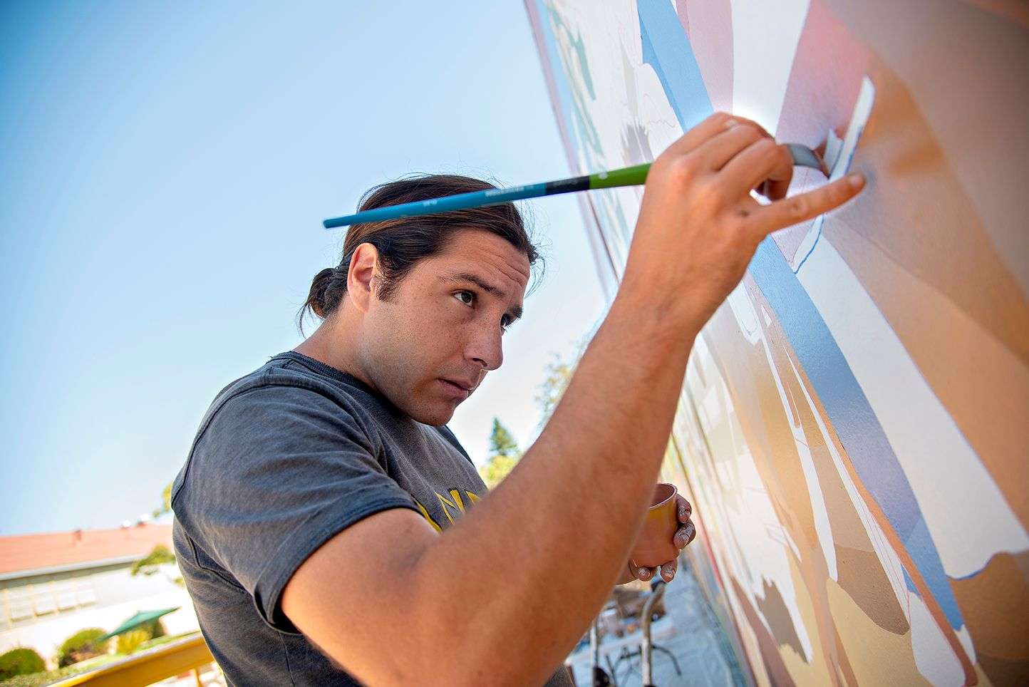 Man with long hair painting a colorful mural on an outdoor wall under clear blue sky.