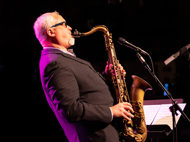 Man with white hair and glasses playing tenor saxophone on stage with microphone and sheet music.